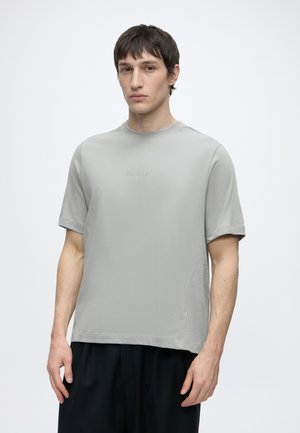 Jeune homme portant un t-shirt gris clair Calvin Klein et un pantalon noir, debout devant un fond blanc uni.