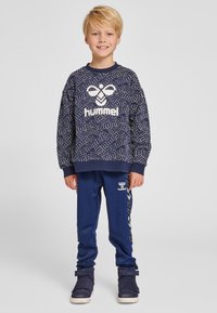 Sweatshirt bleu marine avec un motif géométrique blanc et le logo "hummel" ; assorti avec un jogging bleu marine arborant une marque blanche et des rayures latérales.