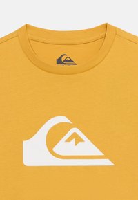 Μουσταρδί κίτρινη μπλούζα με μεγάλο λευκό λογότυπο κύματος και βουνού στο κέντρο κάτω από  το γιακά.