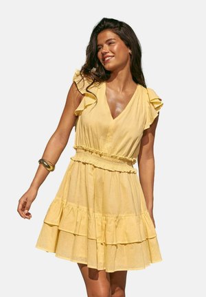 Smilende kvinde med langt hår iført en gul kjole med korte ærmer, flæser og lag samt en guldbangle på højre håndled.