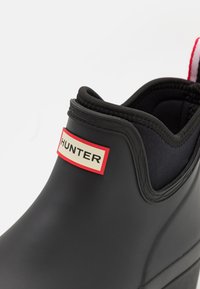 Botte en caoutchouc noire avec un bout arrondi, des côtés élastiques et une étiquette logo rectangulaire en rouge et beige. Texture lisse avec une finition mate.