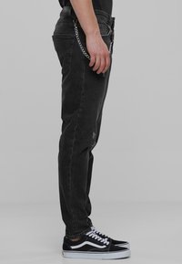 Jeans de denim negro con un corte recto, que presentan una textura desgastada y un acento de cadena plateada en la cinturilla, combinados con zapatillas negras.