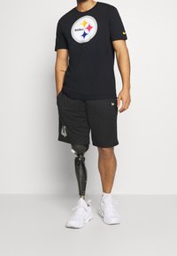 Czarny t-shirt z krótkim rękawem z kolorowym logo Steelers. Uzupełniające czarne szorty z siateczki z detalem logo, w połączeniu z białymi trampkami i protezą nogi.