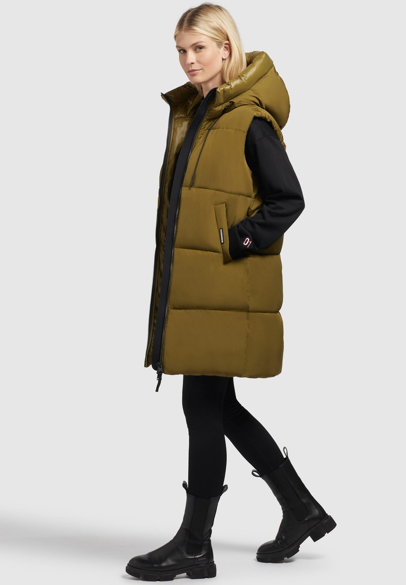 Olivgrüne gesteppte Weste mit Kapuze, Reißverschluss vorne und seitlichen Taschen. Über einem schwarzen Oberteil getragen und kombiniert mit schwarzen Stiefeletten.