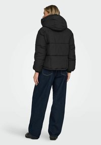 Sort dunjakke med hætte, der har et quiltet design og elastiske manchetter. Bærers over løstsiddende mørke denimbukser, der viser en afslappet pasform.