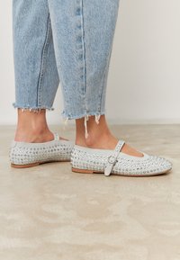 Stopy w jasnoniebieskich balerinkach Mary Jane z ćwiekami i zapięciem, w zestawieniu z dżinsami w jasnoniebieskim kolorze o postrzępionych końcach, na beżowej podłodze.