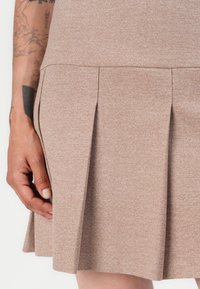 Brown pleated skirt made from a textured fabric, featuring subtle heathered patterns and a smooth hemline. Hand positioned near the fabric.