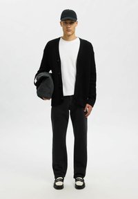 Vyras, dėvintis juodą kepurę, juodą megztinį su užtrauktuku virš balto marškinio, juodas kelnes, laiko pilką sportinį krepšį, stovi prieš paprastą šviesią foną.