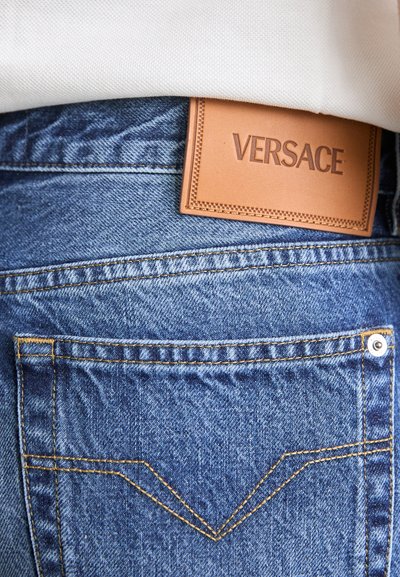 Jeanși din denim albastru cu un buzunar din spate vizibil, decorat cu cusături decorative, completat cu o etichetă din piele bej, gravată cu "VERSACE."