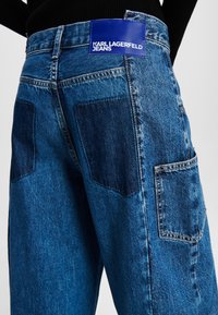 Spijkerbroek met een donkerblauw en lichtblauw patchworkontwerp, met een sterke textuur. Inclusief een blauw logo-label op de taille.
