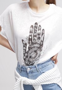 Camiseta de algodón blanca con un gran gráfico de mano negra adornado con varios símbolos y patrones. Combinada con unos vaqueros de denim azul claro y una cinturilla blanca a rayas.