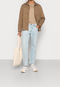 Brown cotton jacket over a beige t-shirt, light blue straight-leg jeans, and beige tote bag. Gray and white sneakers with tan soles.