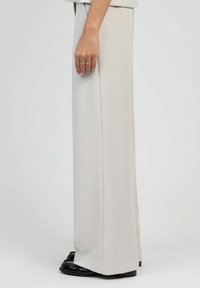 Pantalons larges gris clair avec une texture lisse et douce. Présentent une coupe décontractée et reposent sur des chaussures noires et brillantes.