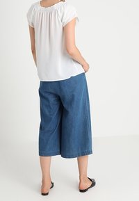 Kvinna i en vit kortärmad blus, bredbeniga blå denimbyxor i caprimodell och svarta slip-in skor, står med händerna i fickorna.