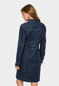 Frau trägt ein dunkelblaues Jeanskleid mit langen Ärmeln, Rückenansicht, stehend vor einem schlichten weißen Hintergrund.