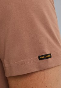 Krátký rukáv, přiléhavé tričko v odstínu starorůžové, vyrobené z bavlny. Na rukávu je černá a žlutá nášivka "PME Legend". Hladká textura.