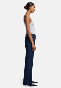 Dunkelblaue Stretch-Denim-Jeans im Bootcut-Stil mit minimalem Verblassen, Fünf-Taschen-Design, sichtbaren Nähten, getragen mit einem hellgrauen Tanktop und schwarzen High Heels.