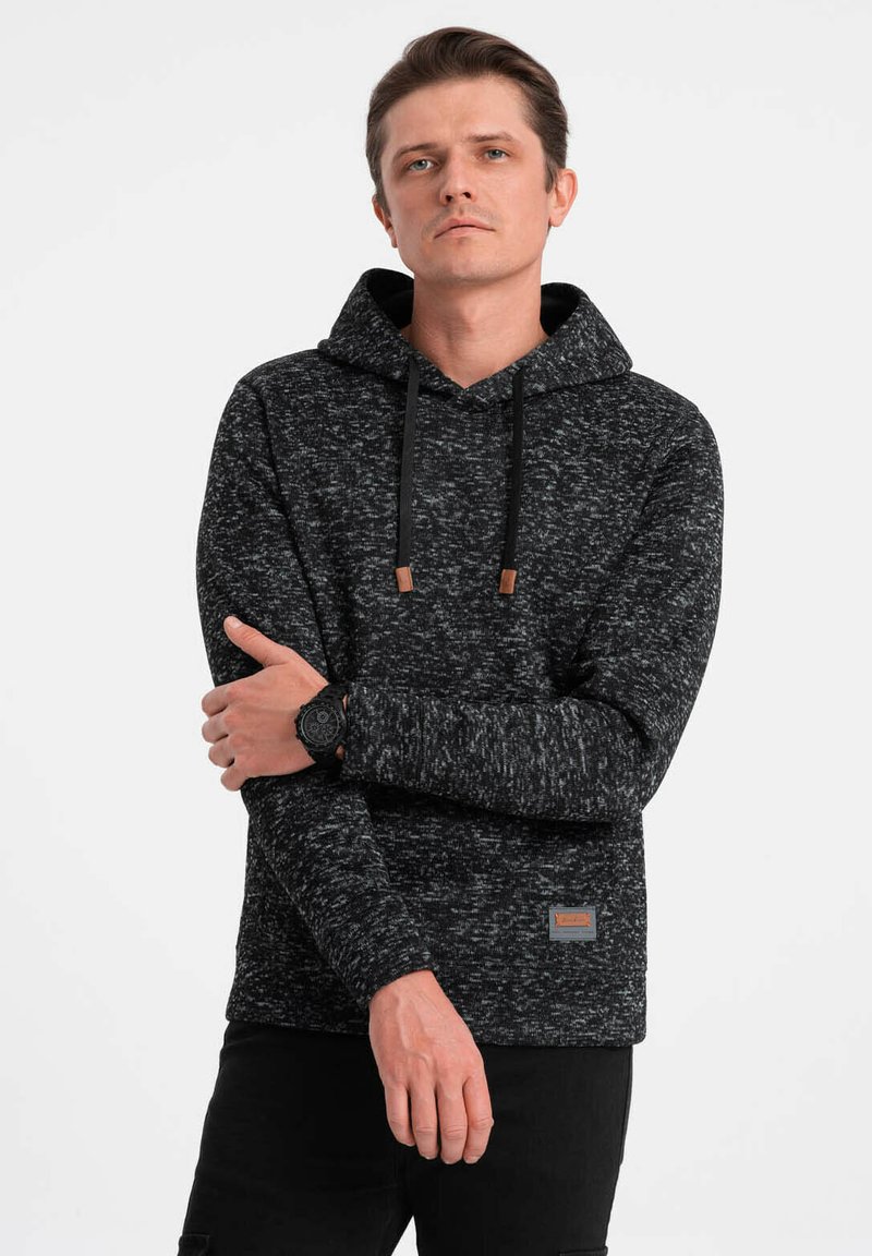 Schwarzer und grauer, gesprenkelter Hoodie mit einer vorderen Tasche, Kordelzugkapuze und Lederakzenten. Das Model trägt dazu schwarze Hosen und eine Uhr.