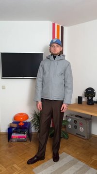 Hallide hallide jope, mis on varustatud ees oleva lukuga, rinnaümbruse tasku ja reguleeritava äärisega, on kombineeritud pruunide pükste ja tumedate kingadega, asetatud tuppa.