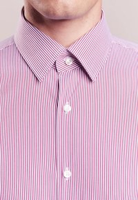 Randig skjorta med tunna vertikala rosa och vita ränder, klassisk krage och vita knappar mot en slät tygtextur.