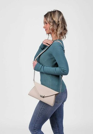 Béžová kožená crossbody taška se stříbrným řetízkovým popruhem, která má trojúhelníkový klapkový design a detail s logem. Nosená s tyrkysovým žebrovaným svetrem.