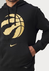 Hombre con sudadera negra con logo de baloncesto dorado y pequeño swoosh de Nike dorado, con la mano en el bolsillo delantero, recortado a la altura del cuello y la cintura.