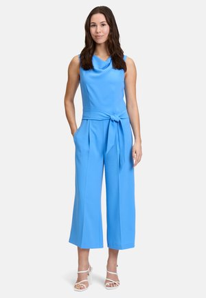 Tuta senza maniche blu con collo drappeggiato, dettaglio in vita annodato, pantaloni a gamba larga e tessuto strutturato. Indossata con tacchi bianchi con cinturini.