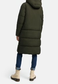 Lång, vadderad kappa i mörkgrön, med huva och horisontell sömnad. Matchad med blå jeans och beige snörstövlar.