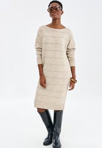 Robe pull en beige clair, ornée de rayures horizontales et de détails en œillets. Manches longues et poignets côtelés. Associée à des bottes noires montant jusqu'aux genoux.