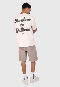 Kremowy t-shirt z krótkim rękawem z napisem "Mindset to Millions" w pogrubionej czarnej czcionce na plecach, zestawiony z beżowymi szortami i białymi sneakersami.