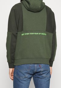 Felpa verde con pannelli a contrasto, caratterizzata da una stampa di testo audace sul retro in verde brillante. Realizzata con un tessuto morbido e strutturato.