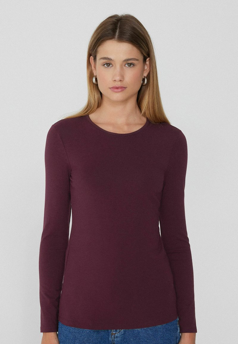 Langärmliges Rundhalsoberteil in tiefem Burgunderrot, aus weichem Material mit glatter Textur. Hat enganliegende Ärmel und ein schlichtes Design.
