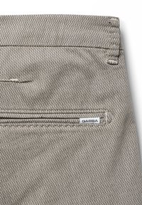 Tejido beige texturizado con un sutil patrón diagonal, que presenta un bolsillo trasero y una pequeña etiqueta blanca "GABBA" cerca del borde del bolsillo.