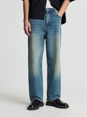 Uomo con jeans a gamba dritta di colore blu chiaro slavato, camicia nera non infilata dentro e mocassini di pelle neri, in piedi con una mano in tasca.