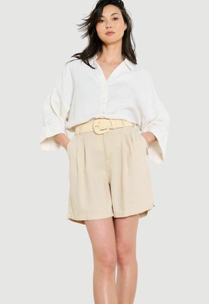 Frau trägt ein locker sitzendes weißes Hemd mit Knöpfen und weiten Ärmeln sowie beige Shorts mit hoher Taille und passendem Gürtel, steht mit den Händen in den Taschen.