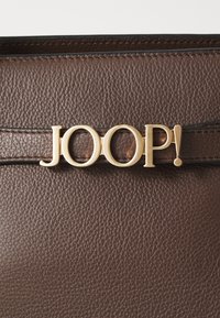 Borsa in pelle marrone con texture e un accento del logo dorato "JOOP!" su una tracolla nera, caratterizzata da cuciture curate e un design elegante.