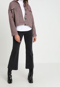 Giacca in houndstooth tagliata in rosso e nero, abbottonata con due tasche sul petto, abbinata a una maglietta bianca e pantaloni neri a campana, stivali lucidi.