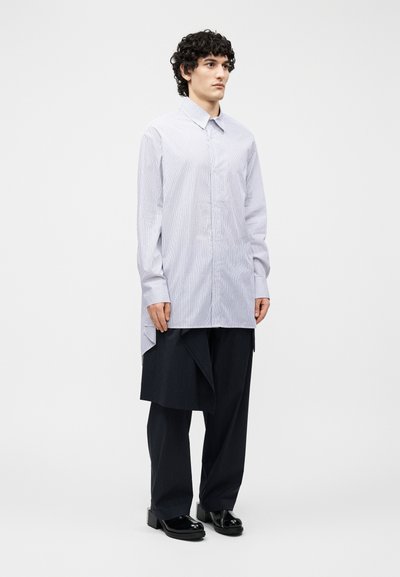 Junger Mann mit lockigem Haar, der ein langes, hell gestreiftes Hemd über schwarzen, weit geschnittenen Nadelstreifenhosen und schwarzen Plateauschuhen trägt.