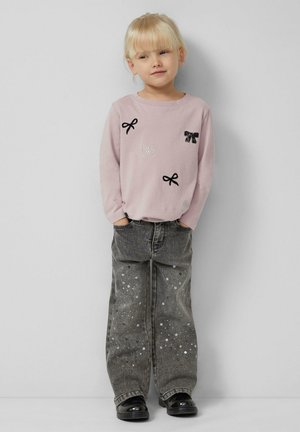 Lichtroze shirt met lange mouwen, versierd met zwarte en zilveren strikken en edelstenen; gecombineerd met losse grijze jeans met paillettenaccenten en zwarte schoenen.