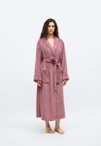 Γυναίκα όρθια, ξυπόλητη, φορώντας μια μακριά, άνετη ρόμπα σε απαλό μωβ χρώμα με τσέπες και ζώνη δεμένη στη μέση, μπροστά σε απλό ανοιχτό φόντο.