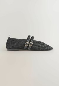 Chaussure plate tissée noire avec bout pointu et deux brides à boucles réglables sur un fond clair.