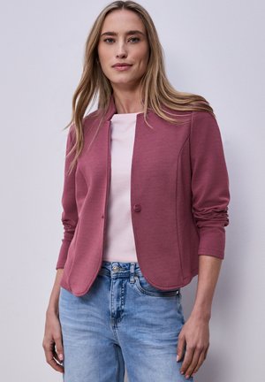 Femme portant un blazer texturé mauve à boutons sur un haut rose clair et un jean bleu clair, debout devant un fond uni.