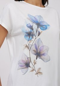 T-shirt en coton blanc avec un imprimé floral dans des tons de bleu et de violet doux, avec des pétales détaillés et un design délicat sur le devant.