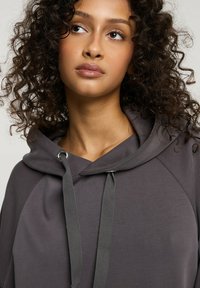 Szary dres z kieszenią na przodzie i kapturem ze sznurkiem. Gładki materiał, luźny krój i rękawy raglanowe zwiększają komfort i styl.