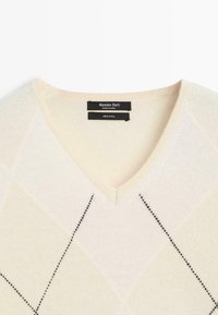 V-hals trui van 100% wol, in crème kleur met een getextureerd ruitpatroon en zwarte stikselaccenten. Merklabel zichtbaar.