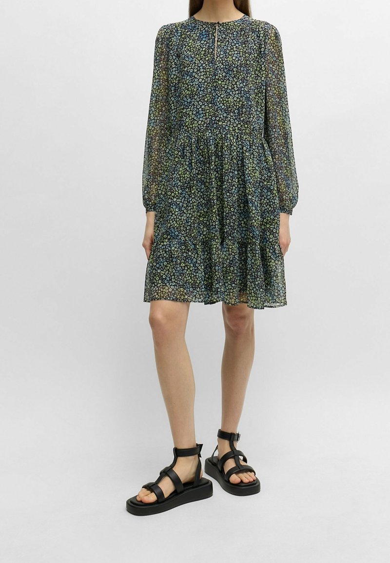 Kleid mit floralem Druck, langen Ärmeln, rundem Ausschnitt und gestuftem Rock. Getragen mit schwarzen Riemchensandalen vor einem einfarbigen Hintergrund.