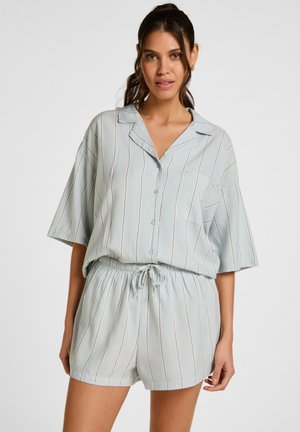 Donna che indossa una camicia button-up a maniche corte a righe verticali azzurro chiaro e pantaloncini coordinati con coulisse, in piedi davanti a uno sfondo grigio uniforme.