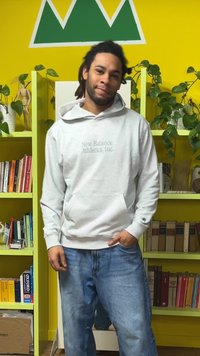 Hanorac gri cu glugă, inscripționat cu "New Balance Athletics, Inc." în gri deschis. Purtat cu blugi largi, de culoare albastru deschis. În fundal se văd rafturi cu cărți și plante.