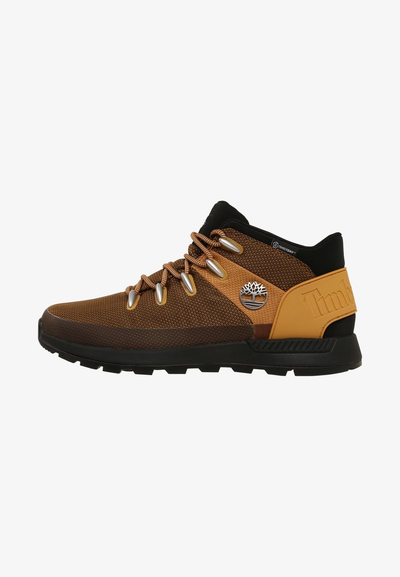 Baskets mi-hautes marron et noires avec tissu texturé, œillets métalliques, logo arbre Timberland sur le côté, et semelle en caoutchouc noir robuste.