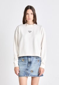 Krémový sveter s krátkym strihom a dlhými rukávmi, s malým logom "Tommy Jeans". V páre s džínsovou mini sukňou, zdobenou okrúhlymi vyšívanými nášivkami.